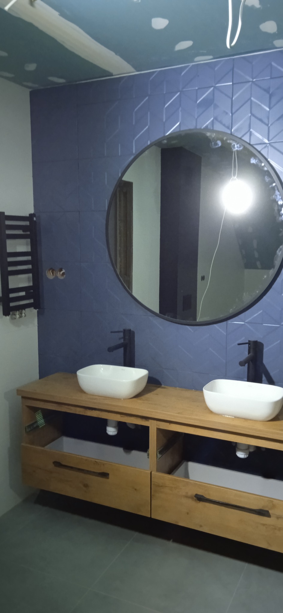 Niebieska łazienka w trakcie wykańczania: dwie białe umywalki nablatowe z czarnymi bateriami, okrągłe lustro w czarnej ramie, grzejnik drabinkowy i ściana z płytek w geometryczny wzór, sufit...