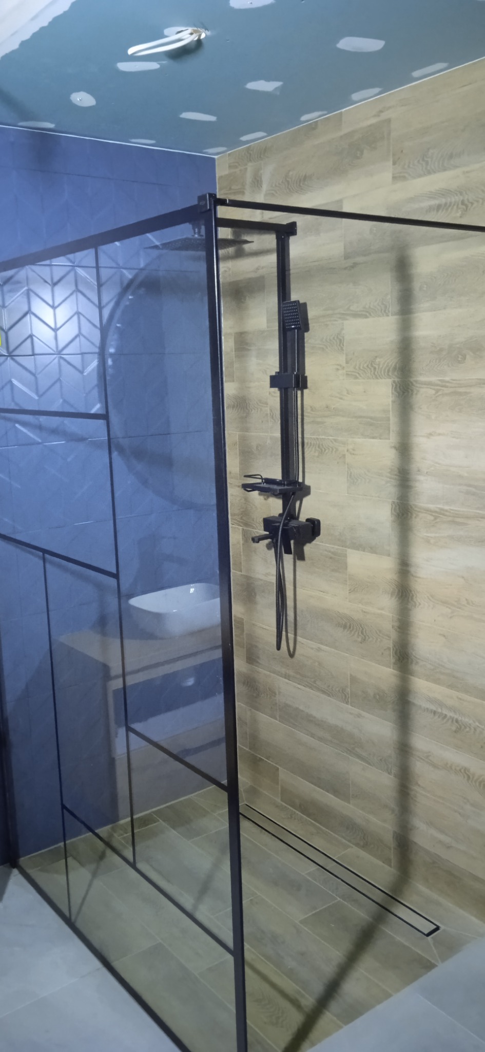 Nowoczesna kabina prysznicowa z czarną armaturą i szklaną ścianką z geometrycznym wzorem, widoczna umywalka nablatowa i niedokończony sufit.