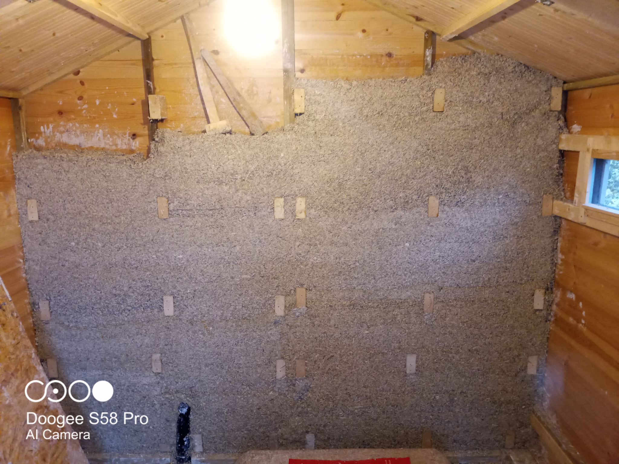 Poddasze domu w trakcie ocieplania, widoczne drewniane elementy konstrukcyjne i warstwa izolacji z wełny celulozowej, przymocowana drewnianymi listwami.