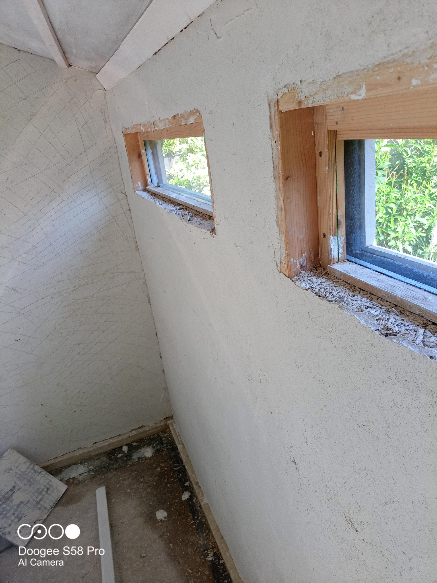 Wnętrze w trakcie remontu z dwoma oknami bez parapetów, ściany z surowym tynkiem z widocznymi śladami po narzędziach, podłoga z resztkami materiałów budowlanych.