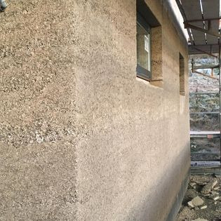 Surowa ściana budynku w trakcie budowy, z widocznym oknem i fragmentem rusztowania, wykończona tynkiem o nierównej fakturze, przypominającym glinę lub ziemię.