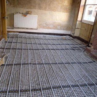 Montaż ogrzewania podłogowego wodnego w remontowanej kamienicy: białe rurki na czarnej siatce, surowe ściany z widocznymi warstwami farby, grzejnik na ścianie, okno w drewnianej ramie.