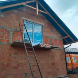 Surowy dom murowany z czerwonej cegły, z zamontowanym oknem, drewnianą drabiną opartą o ścianę i widocznym fragmentem dachu oraz samochodem dostawczym z reklamą okien w tle.