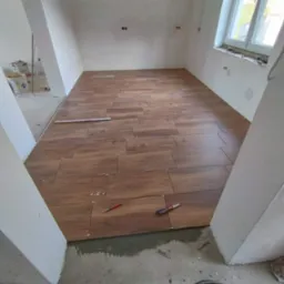 Układanie brązowych płytek imitujących drewno na podłodze w pomieszczeniu w trakcie remontu, widoczne krzyżyki dystansowe, poziomica, n&oacute;ż do cięcia płytek i wiadro z zaprawą.