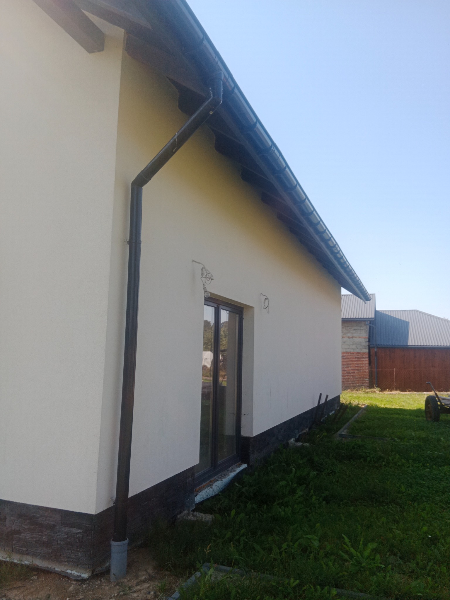 Świeżo otynkowany dom jednorodzinny z ciemnymi rynnami i fragmentem elewacji imitującej kamień u podstawy, widoczne niedokończone podłączenie oświetlenia zewnętrznego.