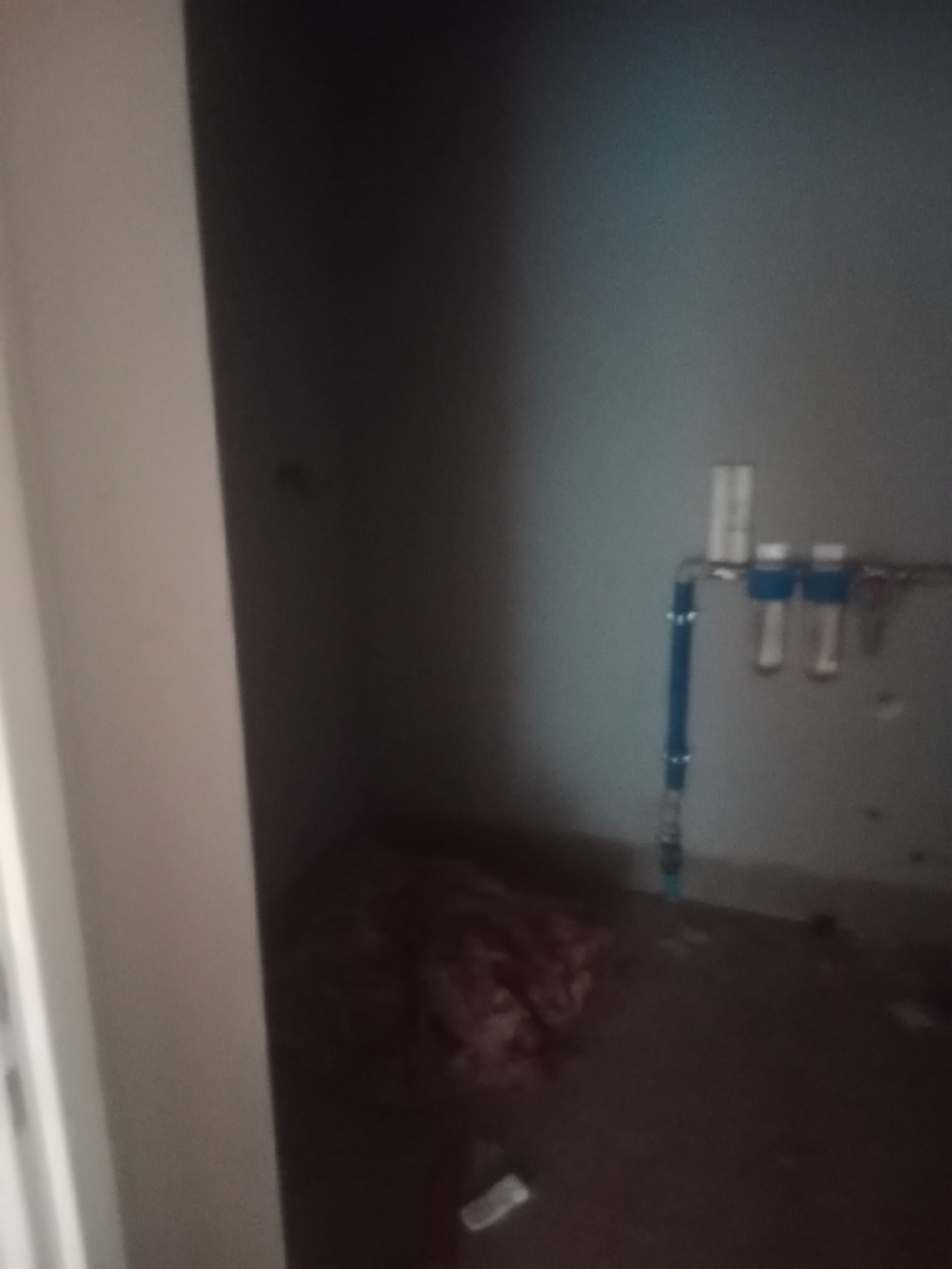 Wnętrze w stanie surowym z instalacją hydrauliczną z niebieskimi rurami i filtrami, widoczne niedociągnięcia w wykończeniu i brak czystości.