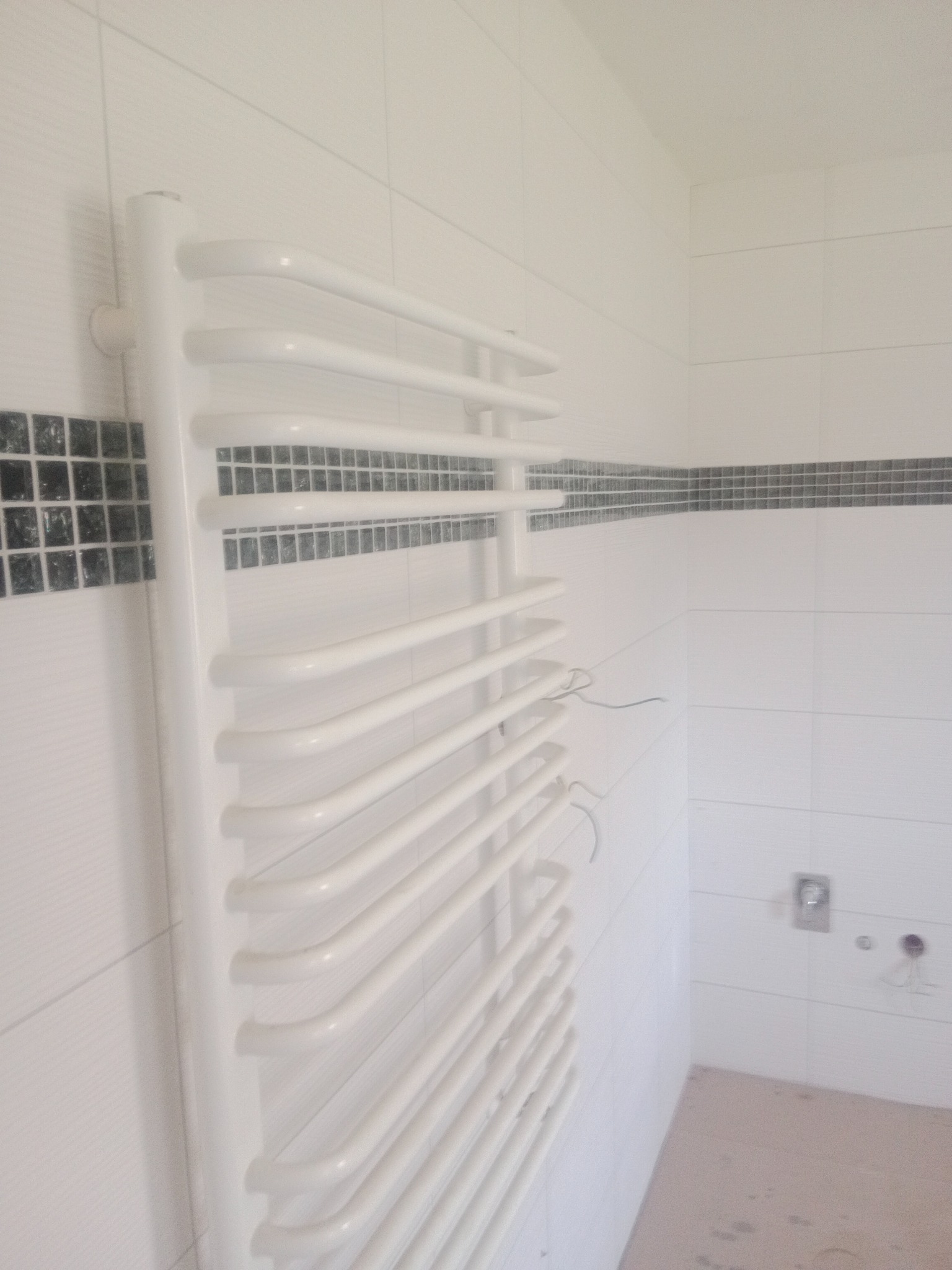Biały, drabinkowy grzejnik łazienkowy zamontowany na białej, wykafelkowanej ścianie z dekoracyjnym pasem czarnej mozaiki. Widoczne wystające przewody instalacji.