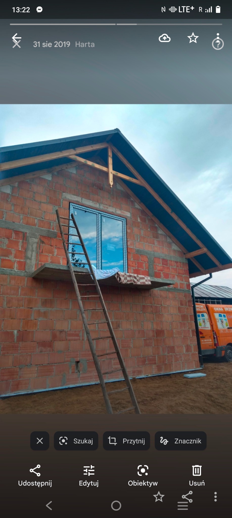 Surowy dom murowany z czerwonej cegły, z zamontowanym oknem, drewnianą drabiną opartą o ścianę i widocznym fragmentem dachu oraz samochodem dostawczym z reklamą okien w tle.