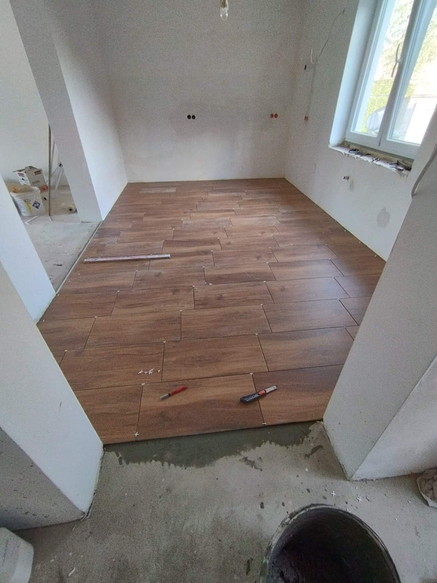 Układanie brązowych płytek imitujących drewno na podłodze w pomieszczeniu w trakcie remontu, widoczne krzyżyki dystansowe, poziomica, nóż do cięcia płytek i wiadro z zaprawą.