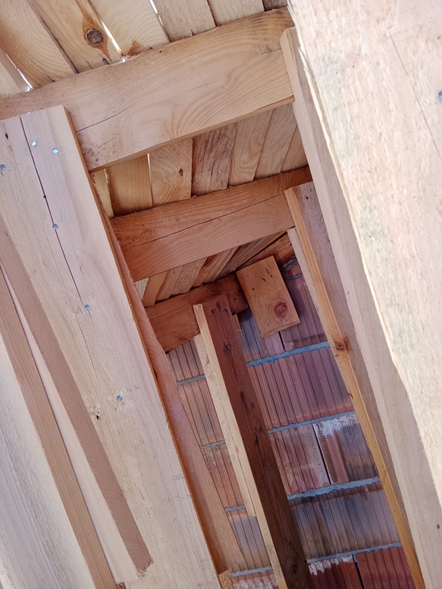 Wnętrze konstrukcji dachu z widoczną drewnianą więźbą i fragmentem murowanej ściany z pustaków ceramicznych, oświetlone naturalnym światłem.