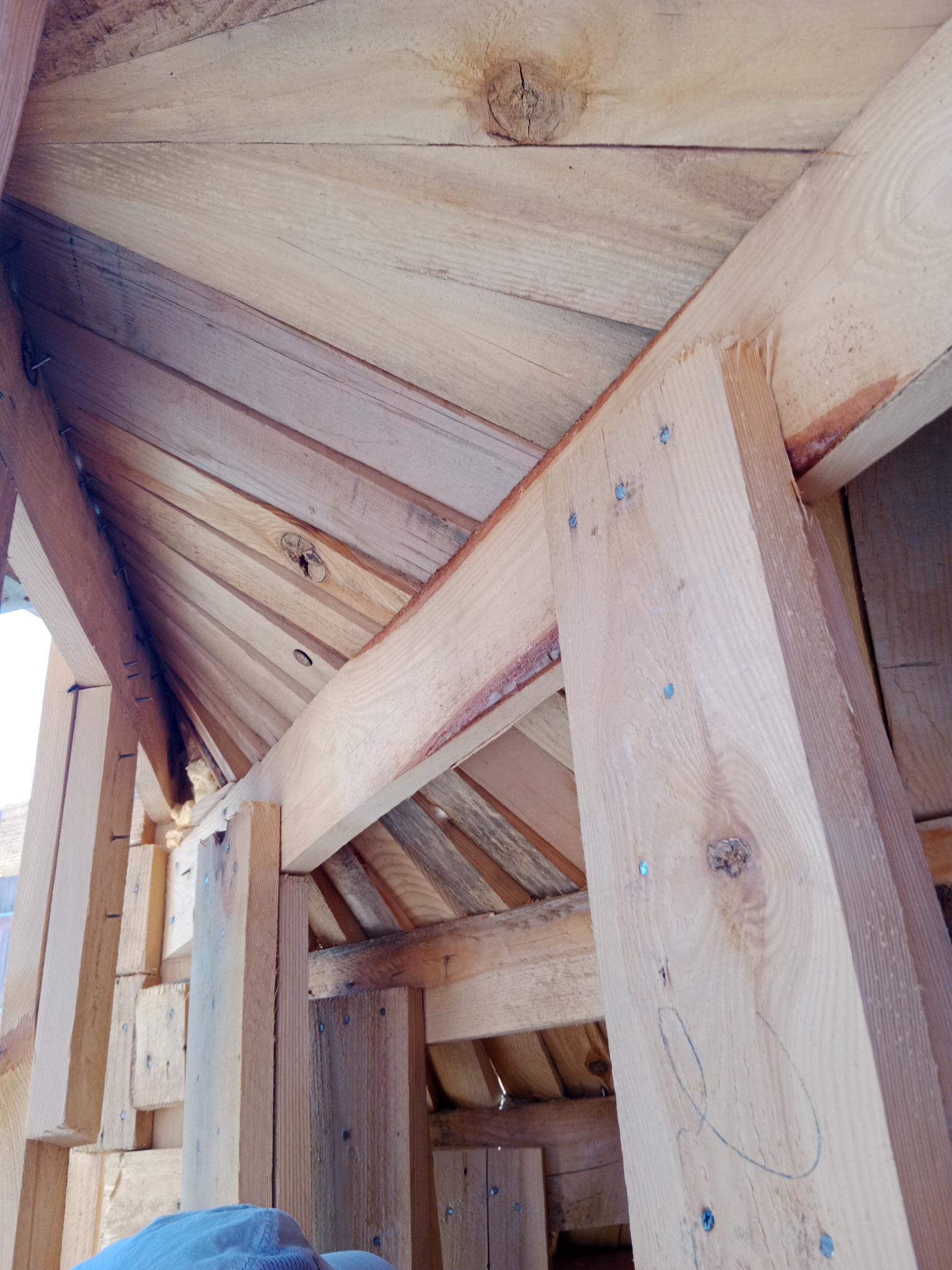 Surowe drewno tworzące konstrukcję dachu, widoczne liczne gwoździe mocujące elementy oraz surowy rysunek na jednym ze słupków.