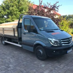 Szary samochód dostawczy z drewnem na pace, zaparkowany na brukowanej nawierzchni. Widoczny budynek w tle i logo firmy na masce.