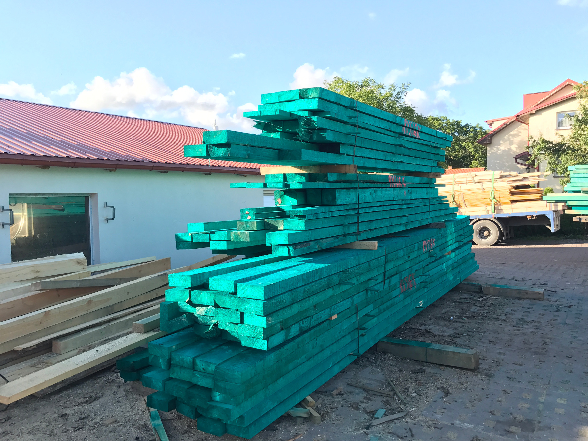Stos impregnowanych, zielonych desek na placu, z widocznym budynkiem i ciężarówką w tle. Selekcjonowane drewno budowlane o wyraźnej strukturze.