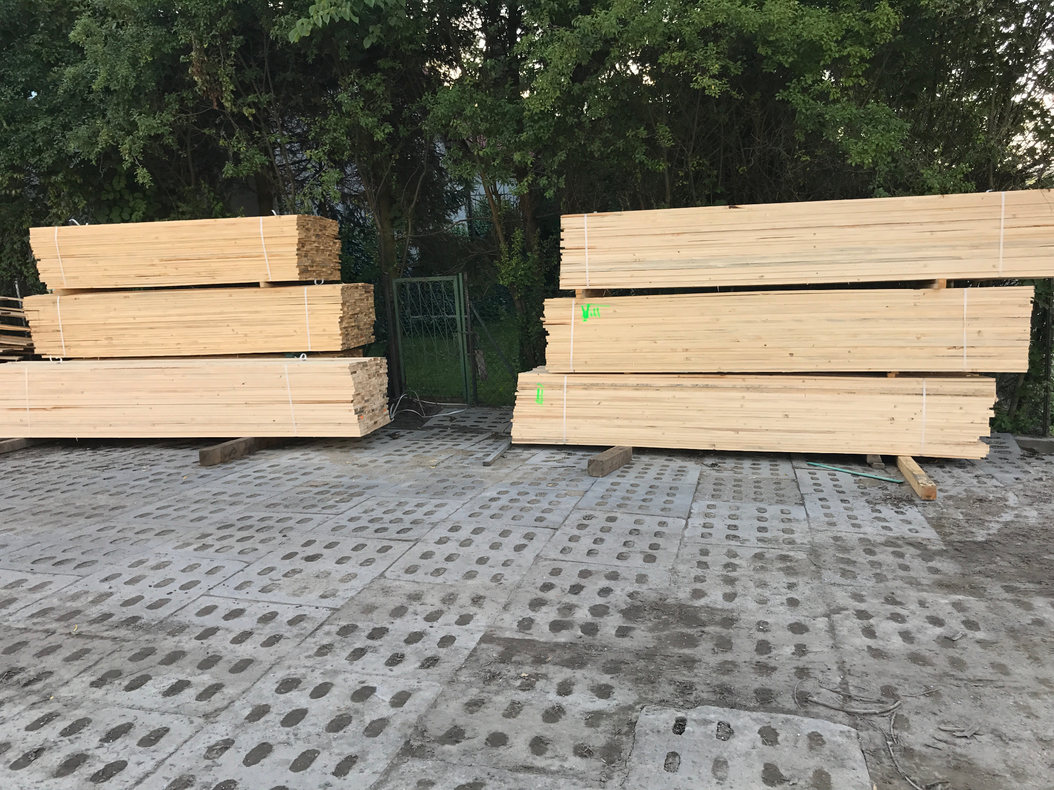 Stosy świeżego drewna na betonowej nawierzchni z zieloną bramką w tle. Widoczne oznaczenia na niektórych belkach.