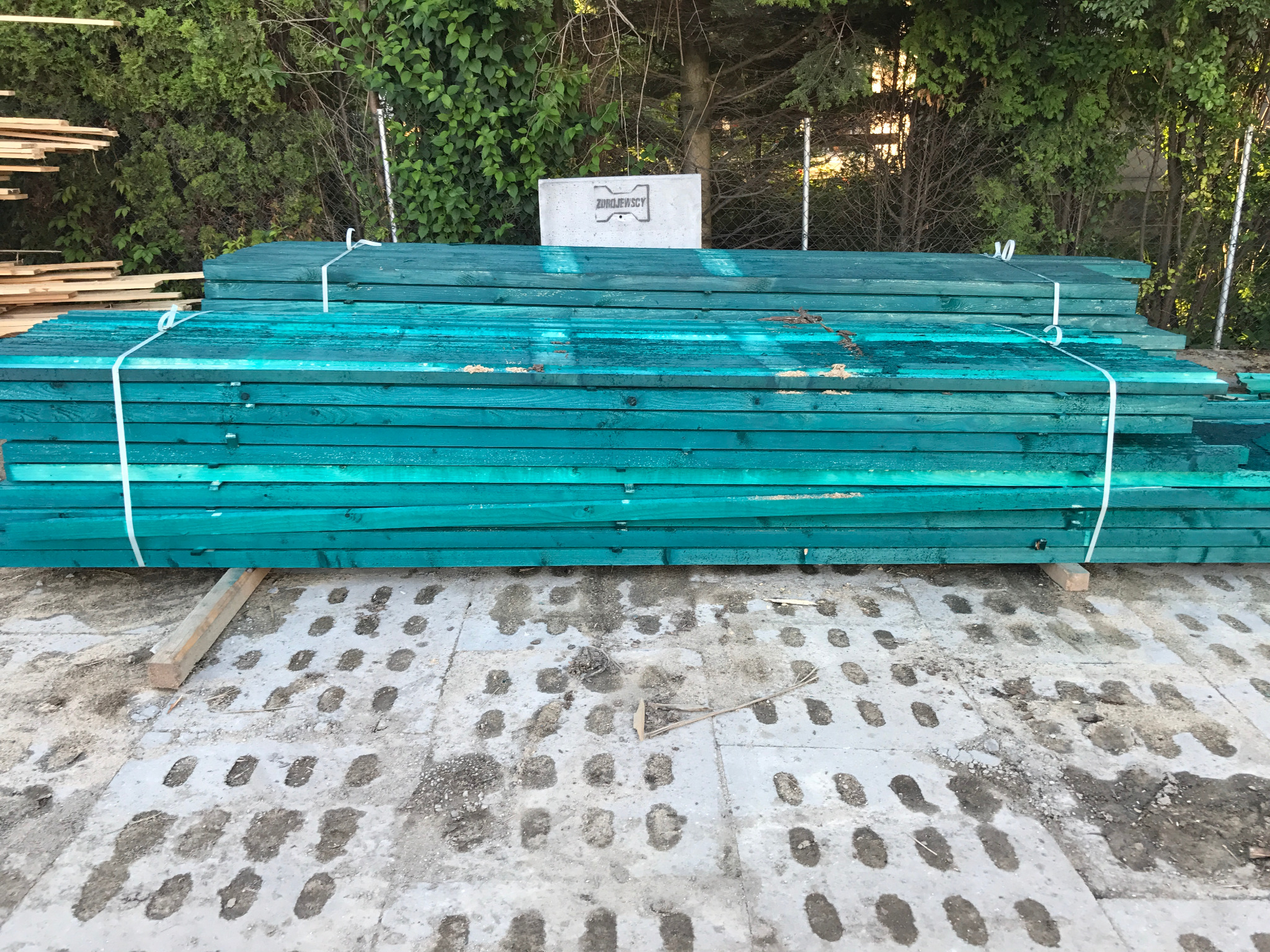 Stos turkusowych desek budowlanych związanych sznurkiem, na betonowej nawierzchni z zielonym tłem roślinnym. Widoczny fragment narzędzia.