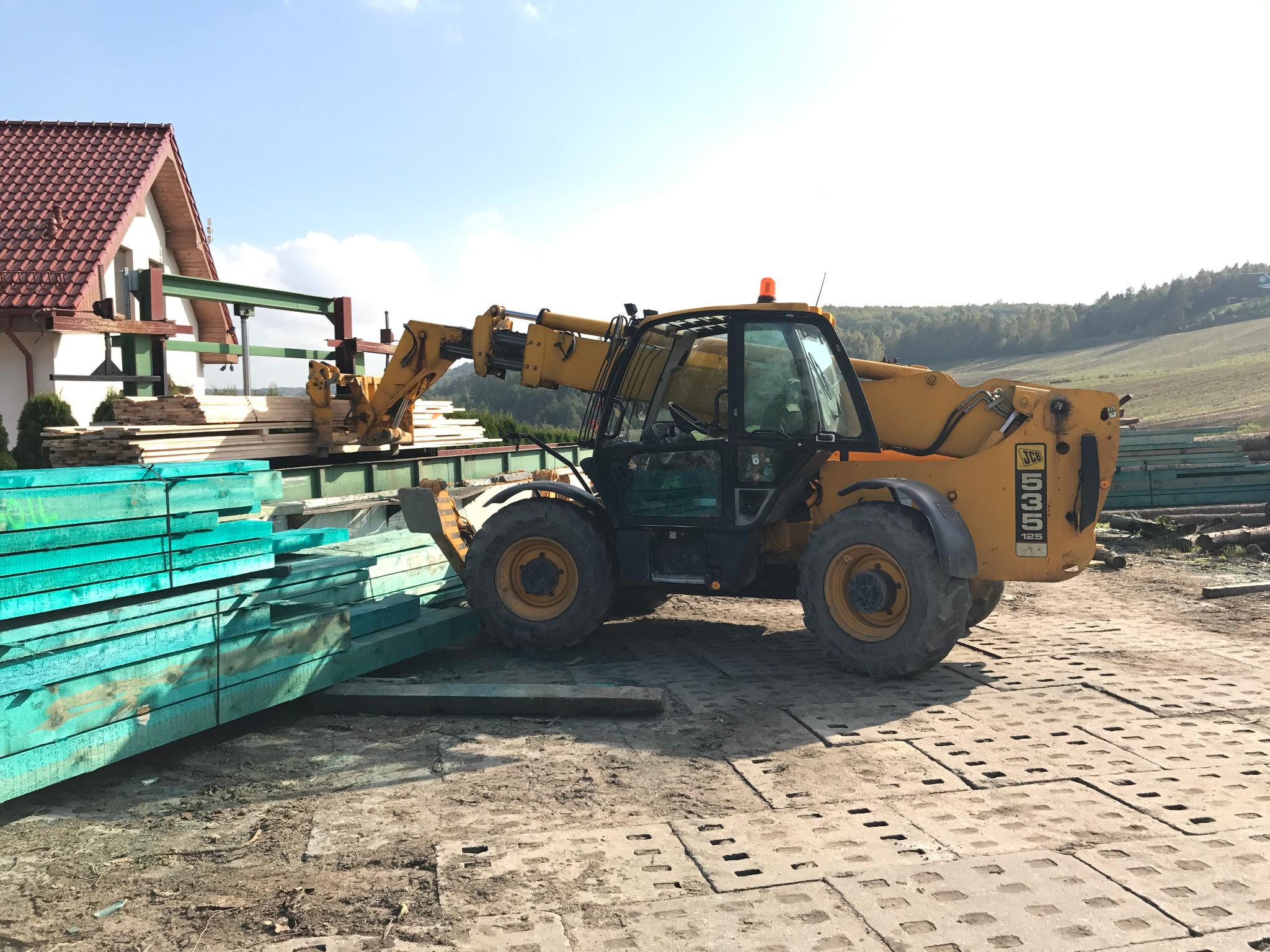 Żółty wózek widłowy JCB podnosi pakiet turkusowych desek na tle budynku i pola w słoneczny dzień. Widoczny stos drewna i betonowa nawierzchnia.