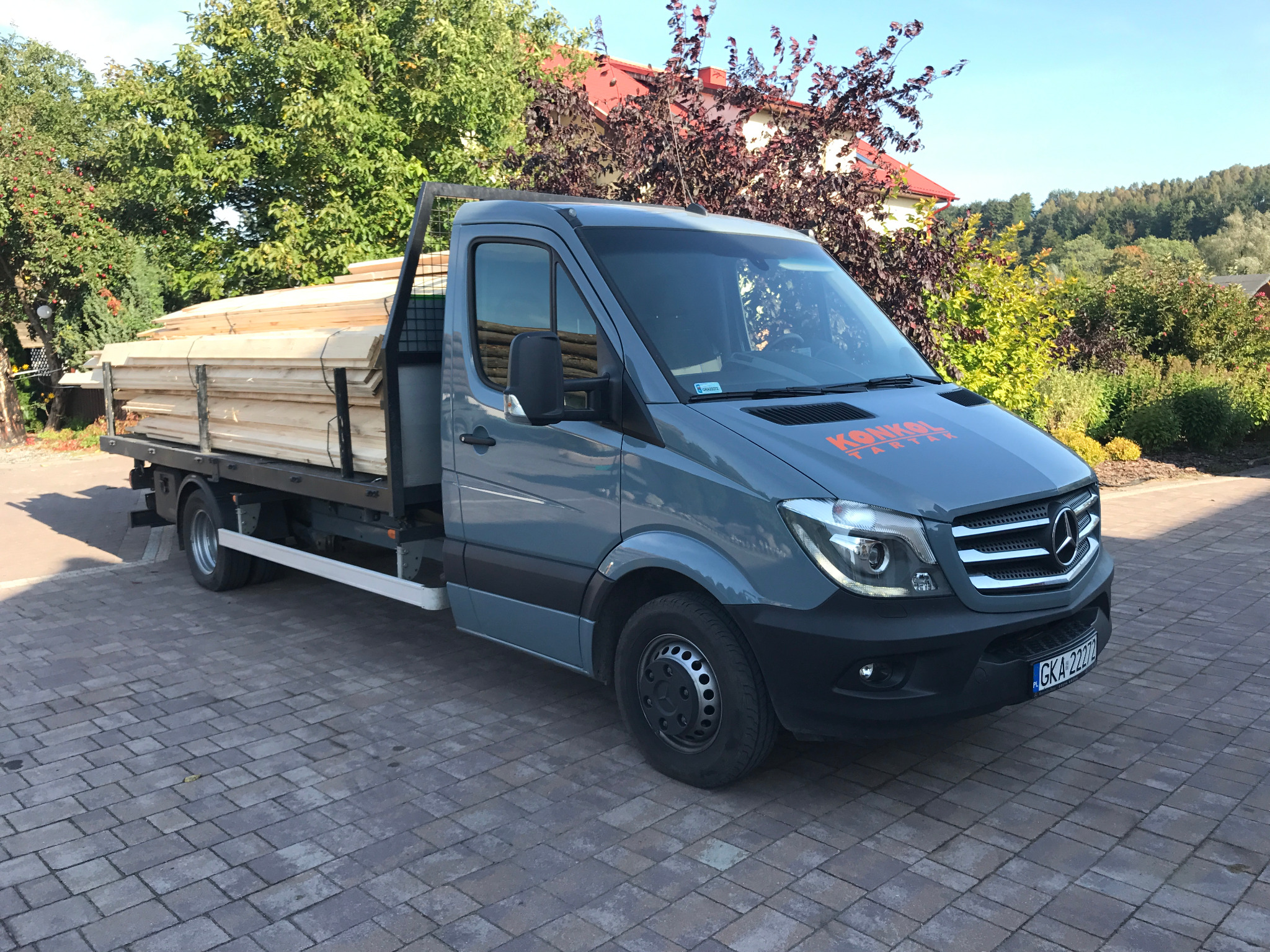 Szary samochód dostawczy z drewnem na pace, zaparkowany na brukowanej nawierzchni. Widoczny budynek w tle i logo firmy na masce.