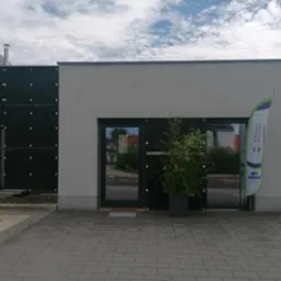 Front budynku z ciemnymi drzwiami i oknami, przed wejściem roślina w donicy, obok flaga reklamowa z napisem 'Ekologia kosmetyki zdrowe kosmetyki'.