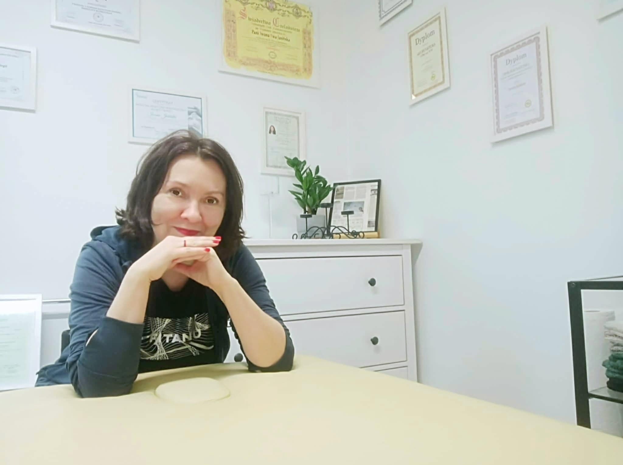 Kobieta z czerwonymi paznokciami siedzi przy stole do masażu w gabinecie z dyplomami na ścianie i komodą w tle. Uśmiech, profesjonalizm, ciepłe oświetlenie.