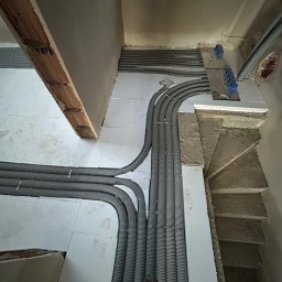 Usługi Hydrauliczne Bartłomiej Leja - Szare, karbowane rury instalacyjne ułożone równolegle na podłodze z jasnych płytek, prowadzące do rozdzielaczy z niebieskimi elementami, widoczne schody w tle.