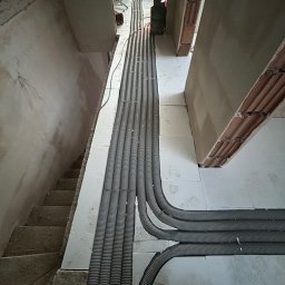 Usługi Hydrauliczne Bartłomiej Leja - Szare, karbowane rury wentylacyjne ułożone równolegle na białych płytach izolacyjnych w surowym wnętrzu budynku w trakcie budowy, widok z góry, fragment schodów i butów roboczych w kadrze.