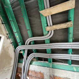 Usługi Hydrauliczne Bartłomiej Leja - Szare, karbowane rury wentylacyjne ułożone równolegle pod skośnym dachem z zielonymi belkami konstrukcyjnymi i czarną membraną, widoczne otwory w murze z cegły.