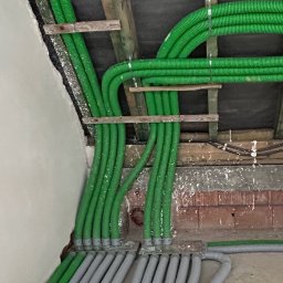 Usługi Hydrauliczne Bartłomiej Leja - Zielone i szare karbowane rury wentylacyjne zamontowane pod sufitem, przymocowane do drewnianych belek, widoczny fragment ściany i podłogi.