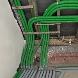 Zielone i szare karbowane rury wentylacyjne zamontowane pod sufitem, przymocowane do drewnianych belek, widoczny fragment ściany i podłogi.