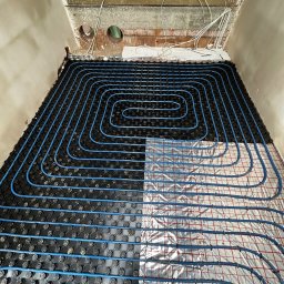 Usługi Hydrauliczne Bartłomiej Leja - Niedokończona instalacja ogrzewania podłogowego: niebieskie rurki ułożone na czarnych mocowaniach, częściowo na izolacji z siatką, widoczne ściany w surowym stanie i drewniany strop z instalacją...