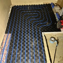 Usługi Hydrauliczne Bartłomiej Leja - Montaż ogrzewania podłogowego w Czarnym Dunajcu: niebieskie rurki ułożone na czarnej płycie systemowej z folią aluminiową, widoczne narzędzia i materiały montażowe.