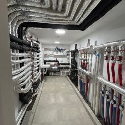 Usługi Hydrauliczne Bartłomiej Leja - Węzeł cieplny z rzędami izolowanych rur w kolorze srebrnym i czarnym, prowadzących wzdłuż ścian i sufitu, z licznymi zaworami, manometrami i przewodami w kolorach czerwonym i niebieskim...