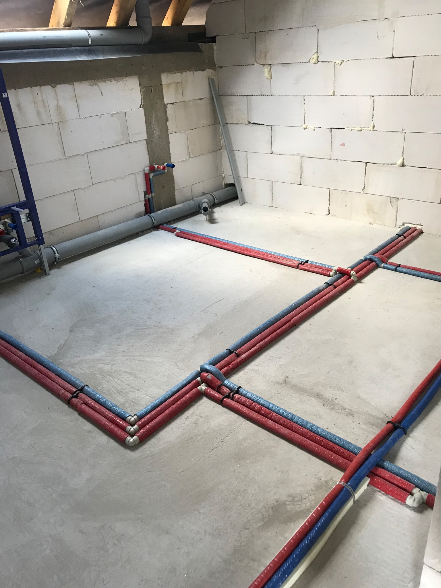 Instalacja wodna podłogowa w surowym stanie, rury czerwone i niebieskie ułożone na szarym betonie, ściany z białego gazobetonu, widoczny fragment stelaża podtynkowego.