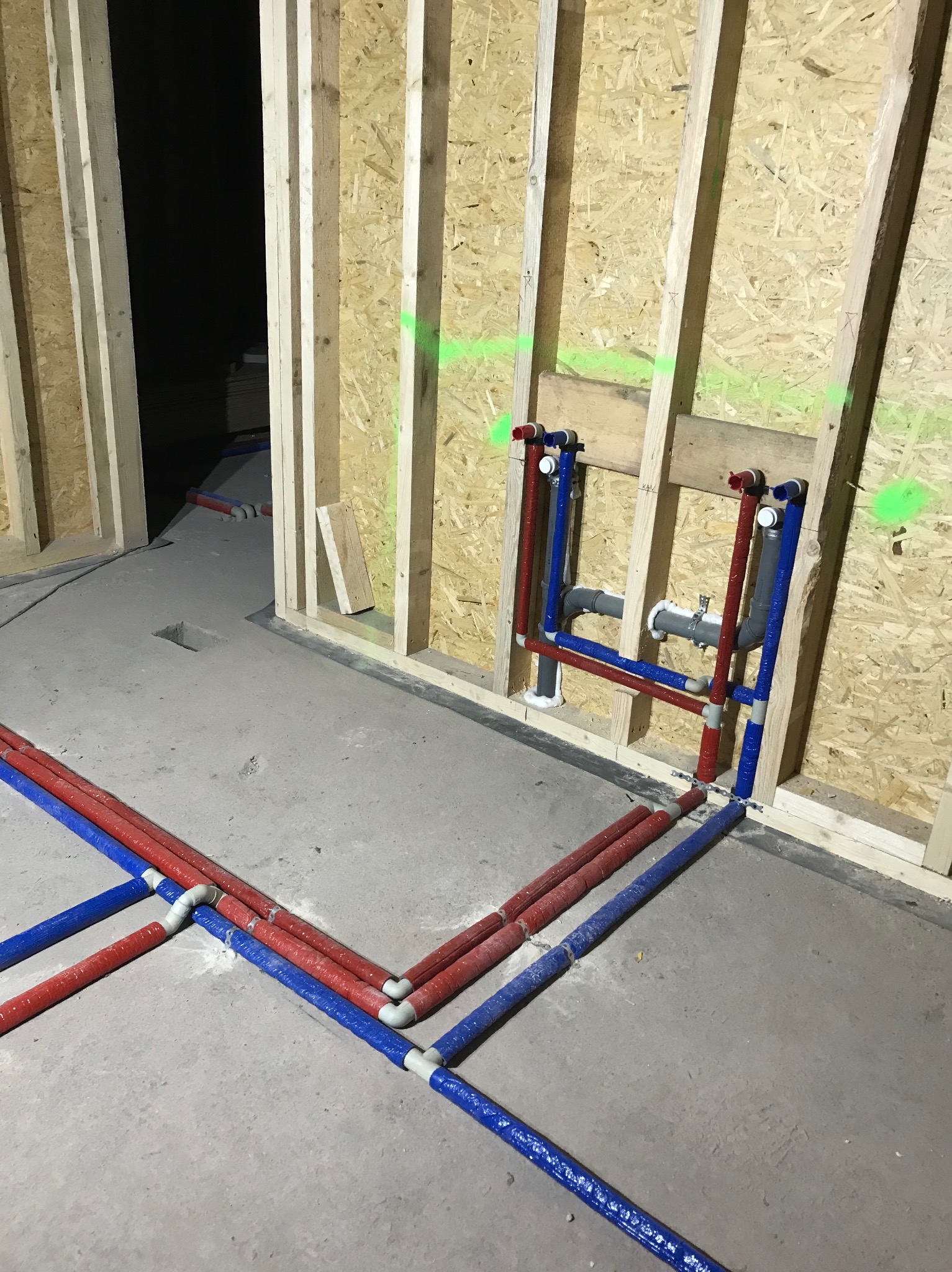 Instalacja wodna PEX w budynku w trakcie budowy, rury czerwone i niebieskie prowadzone po betonowej posadzce i zamocowane do drewnianej ściany szkieletowej, widoczne złącza i izolacja.