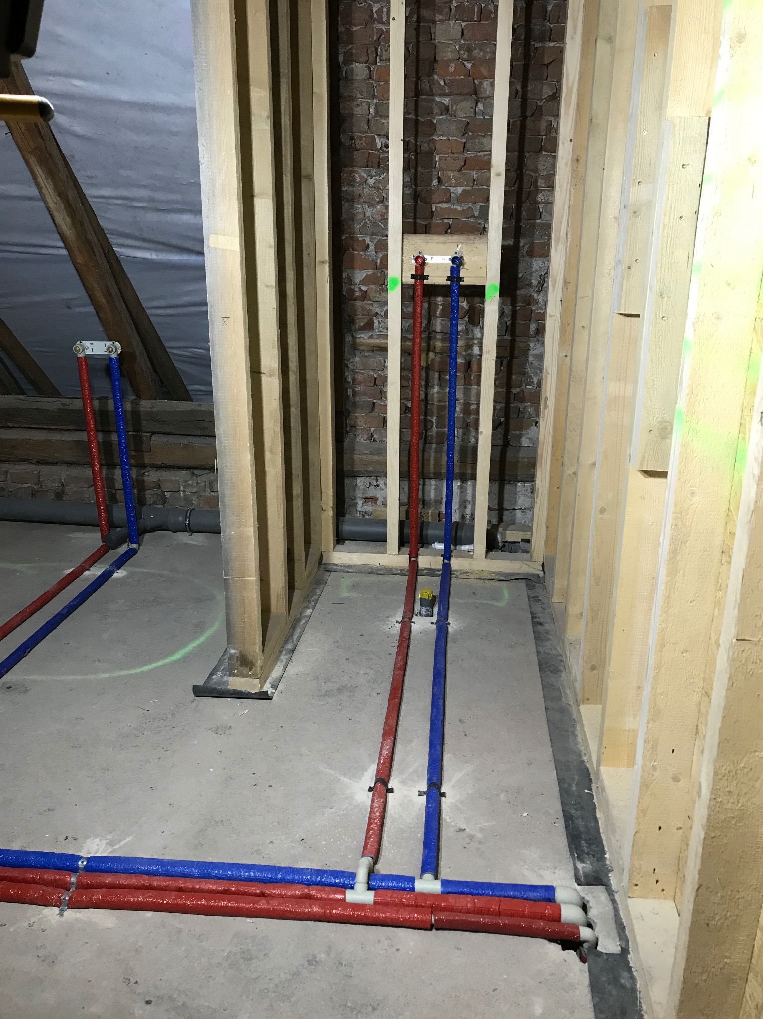 Nowa instalacja wodna z rur PEX w kolorze czerwonym i niebieskim, prowadzone wzdłuż drewnianej konstrukcji szkieletowej i ściany z czerwonej cegły w surowym stanie.