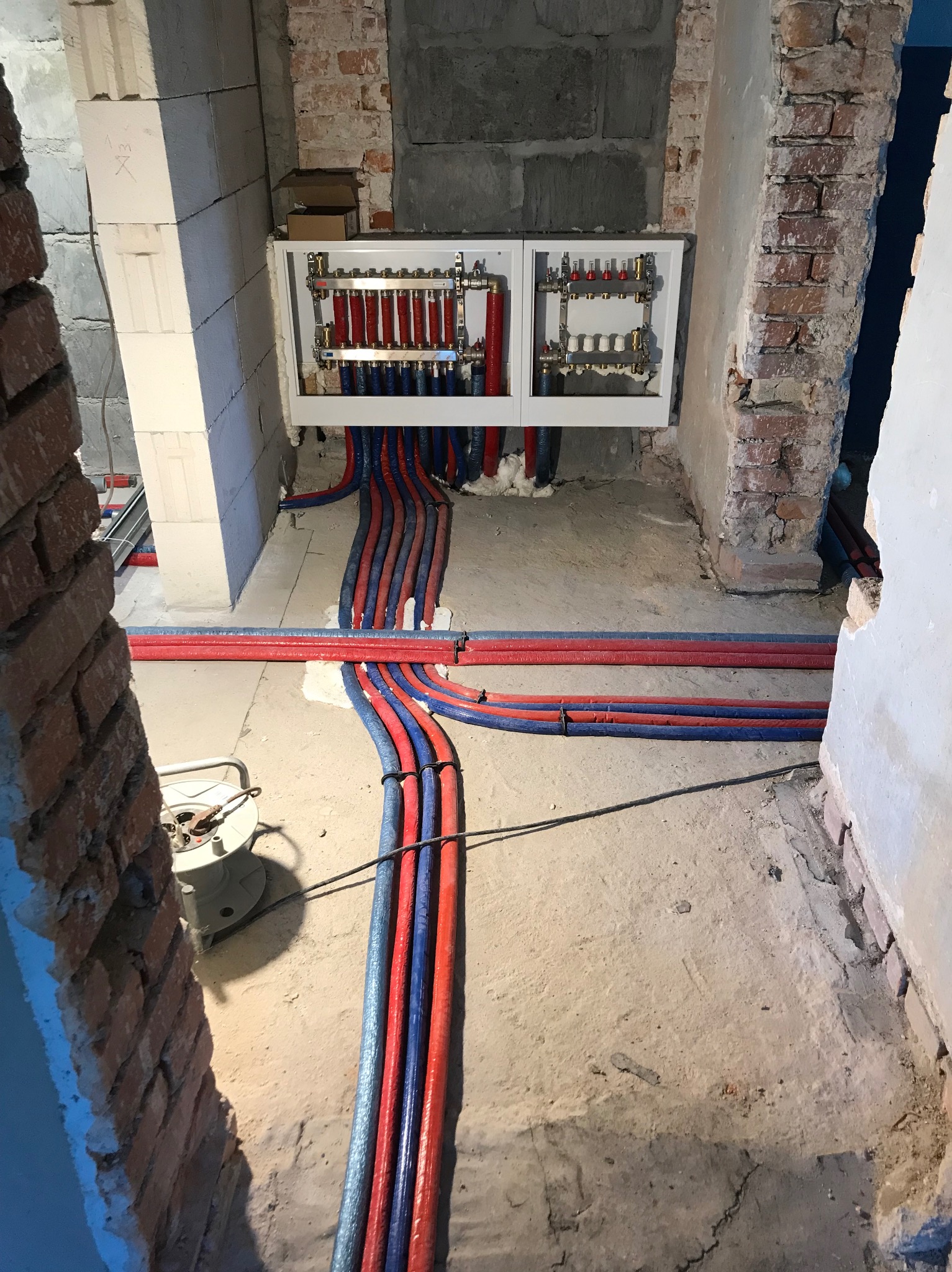 Rozdzielacz instalacji grzewczej podtynkowej z rurami w kolorze czerwonym i niebieskim, na surowym betonie w trakcie montażu w pomieszczeniu z odsłoniętą cegłą.