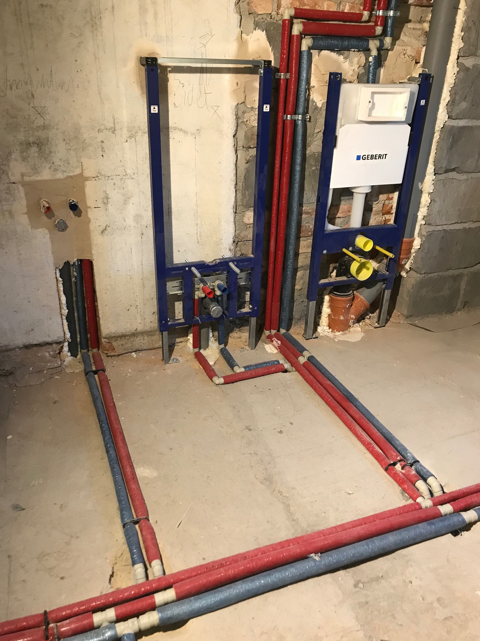 Instalacja podtynkowa Geberit z rurami w izolacji czerwonej i niebieskiej, widoczne mocowania do ściany w surowym stanie.