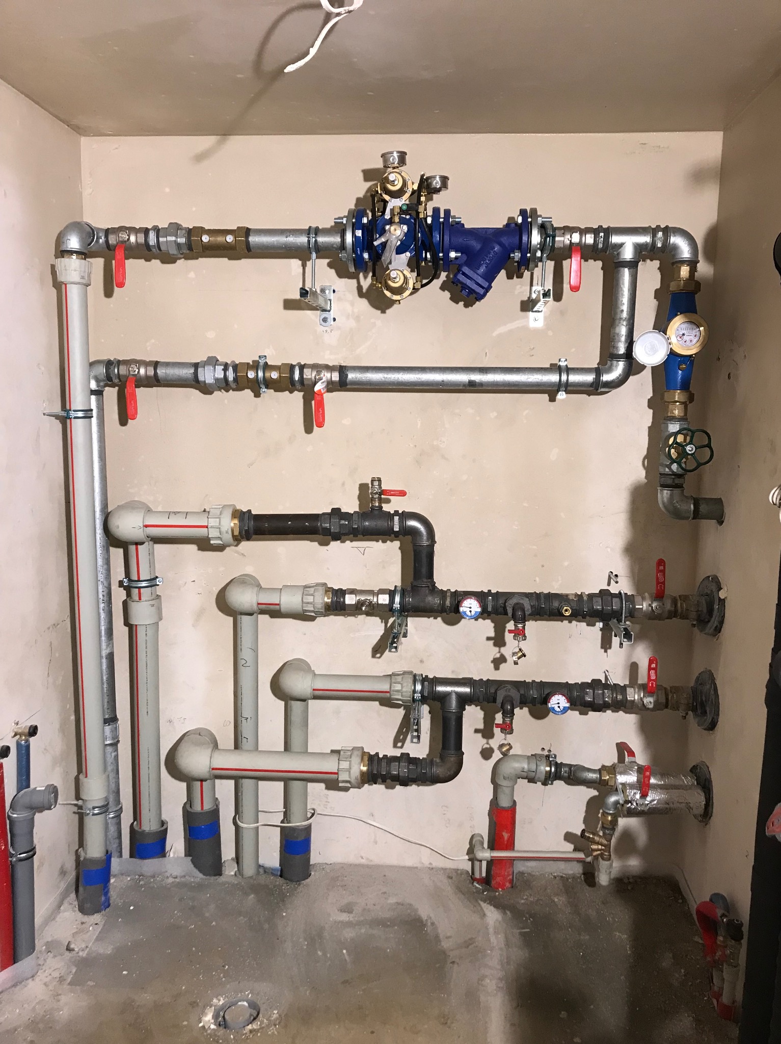 Skomplikowana instalacja wodna z rur stalowych, miedzianych i plastikowych, zaworami z czerwonymi rączkami, manometrami i filtrem w kolorze niebieskim, zamontowana na surowej ścianie w trakcie...