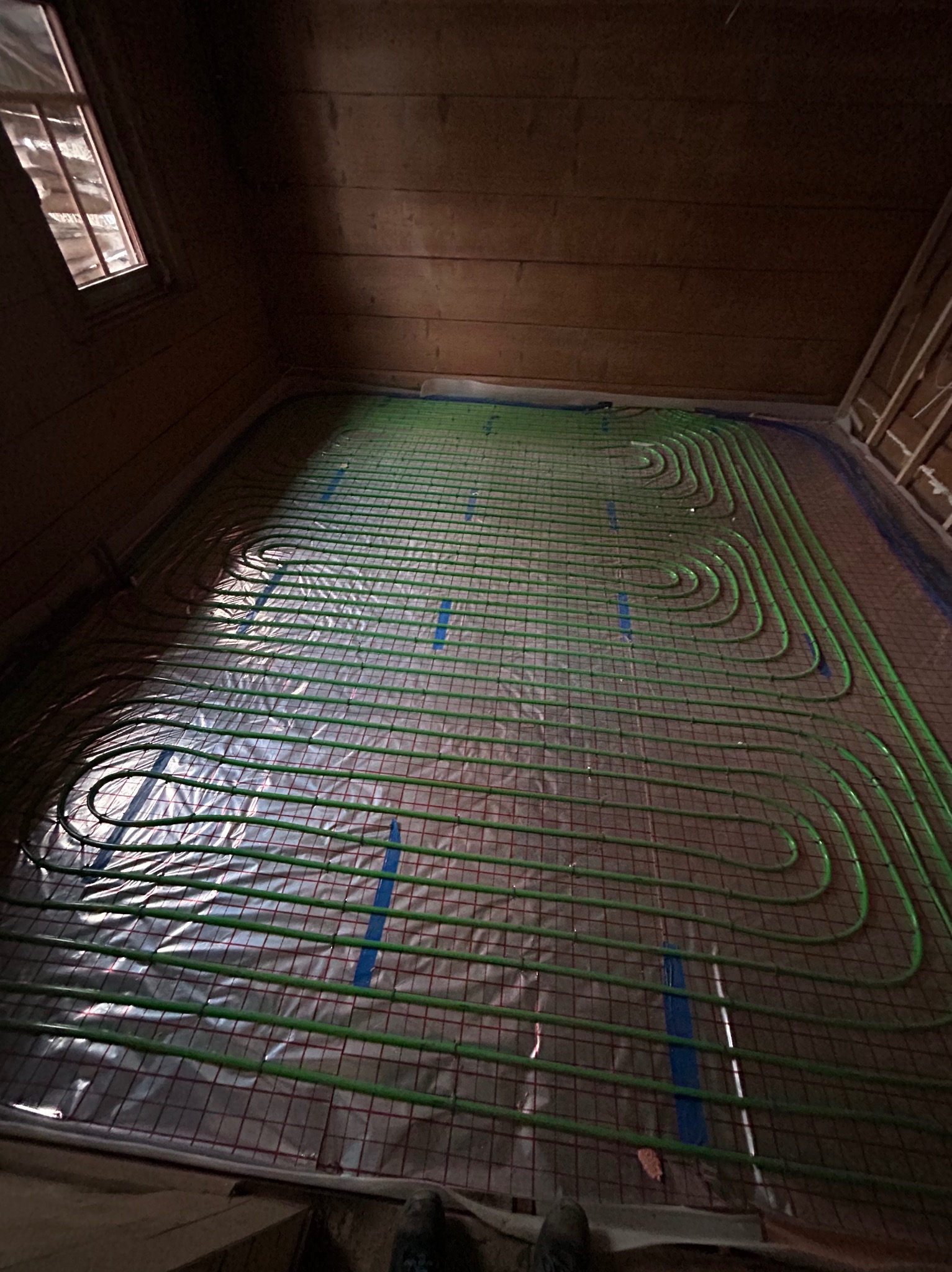 Zielone rurki instalacji grzewczej podłogowej ułożone na folii z siatką w drewnianym pomieszczeniu, widoczne fragmenty butów instalatora w dolnej części kadru.