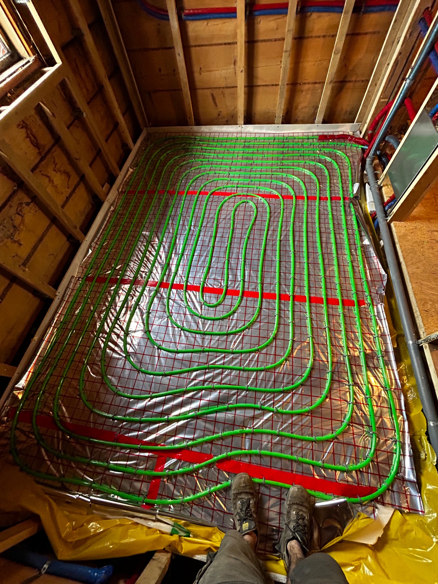 Montaż ogrzewania podłogowego z zielonych rurek na folii refleksyjnej w drewnianej konstrukcji poddasza, widoczne buty montera w dolnej części kadru.