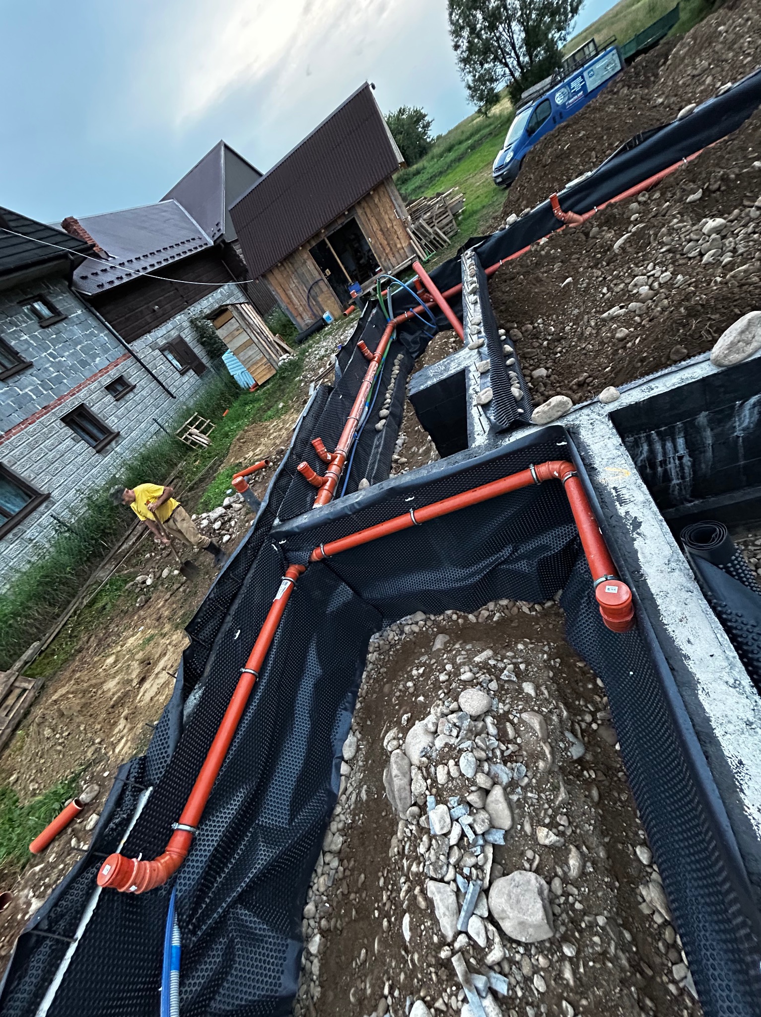 Fundamenty budynku z zainstalowaną izolacją przeciwwilgociową i częściowo ułożonymi rurami kanalizacyjnymi w kolorze pomarańczowym, widoczny pracownik w żółtej koszulce i budynki w tle.