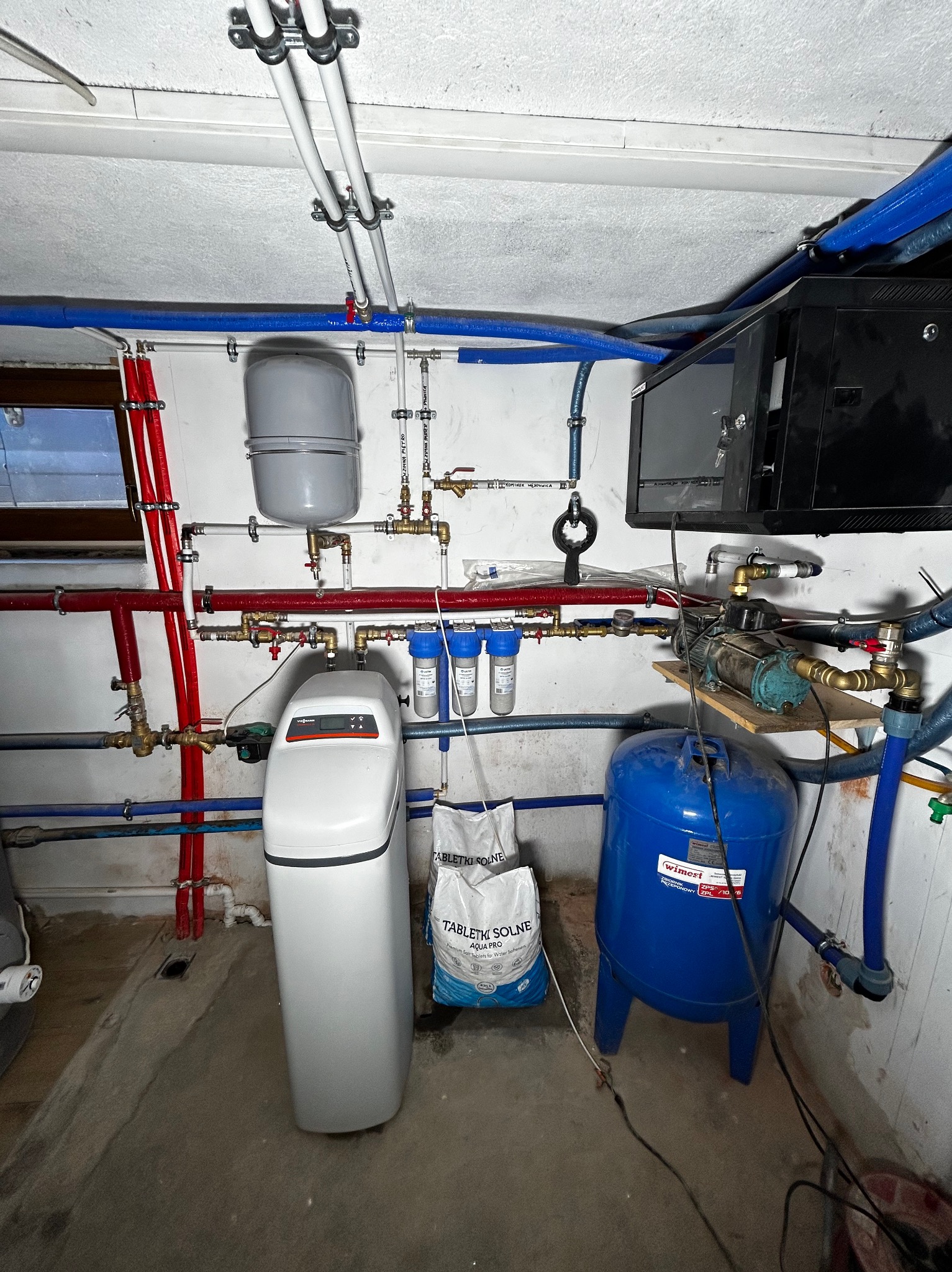 Instalacja wodna w piwnicy: zmiękczacz wody Viessmann, niebieski zbiornik ciśnieniowy Wimest, filtry mechaniczne, liczne rury miedziane i plastikowe, zawory oraz szafka sterownicza na ścianie.