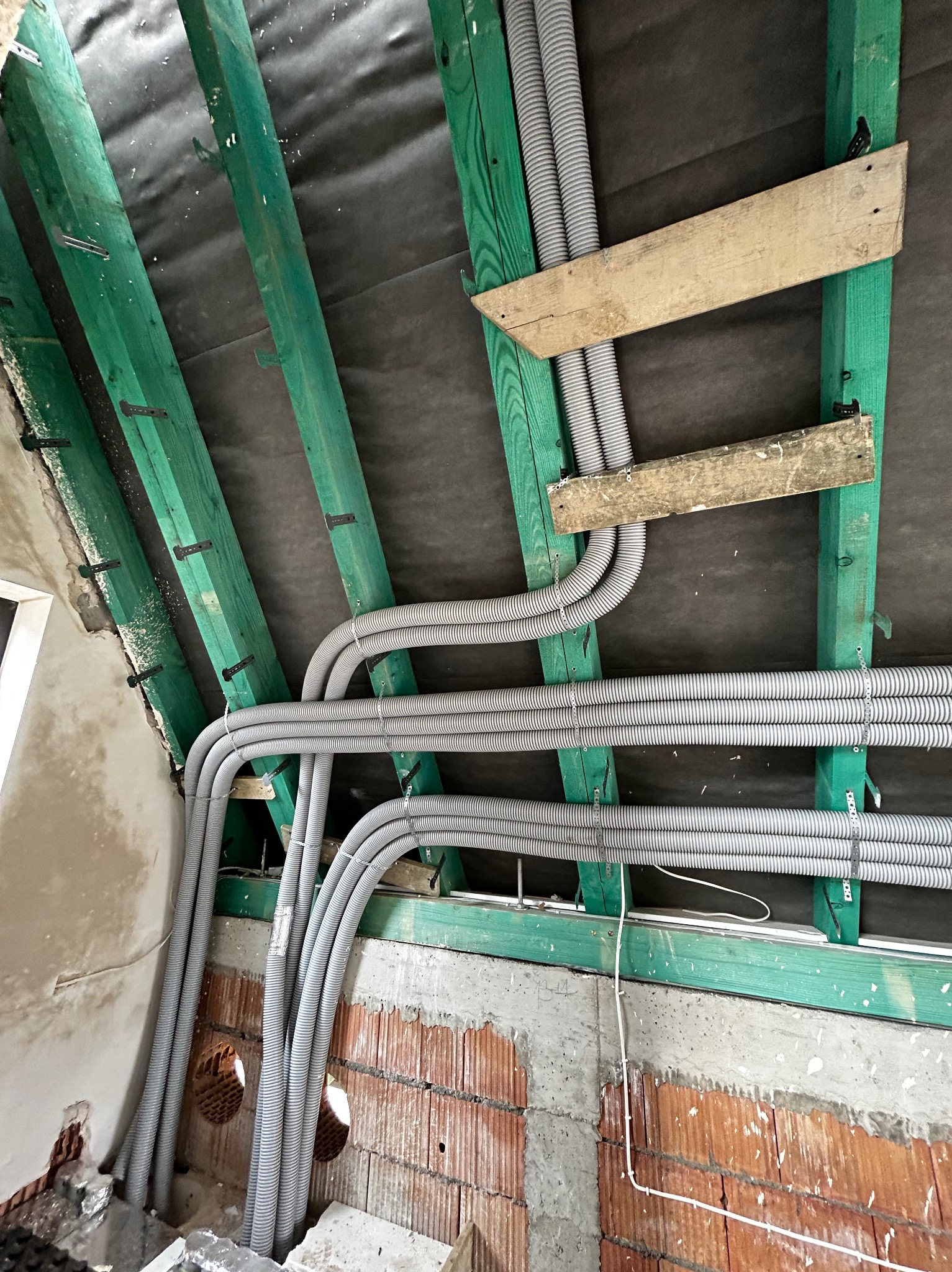 Szare, karbowane rury wentylacyjne ułożone równolegle pod skośnym dachem z zielonymi belkami konstrukcyjnymi i czarną membraną, widoczne otwory w murze z cegły.