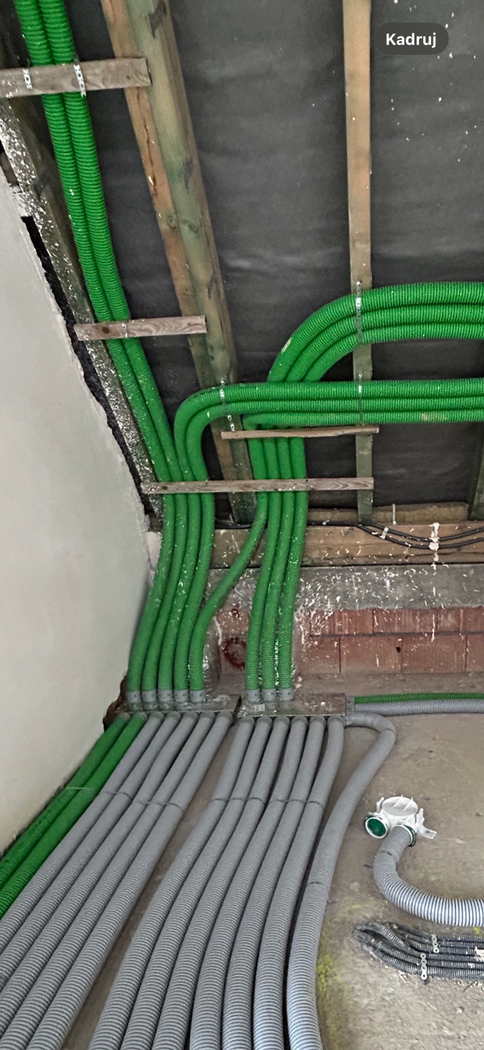 Zielone i szare karbowane rury wentylacyjne zamontowane pod sufitem, przymocowane do drewnianych belek, widoczny fragment ściany i podłogi.