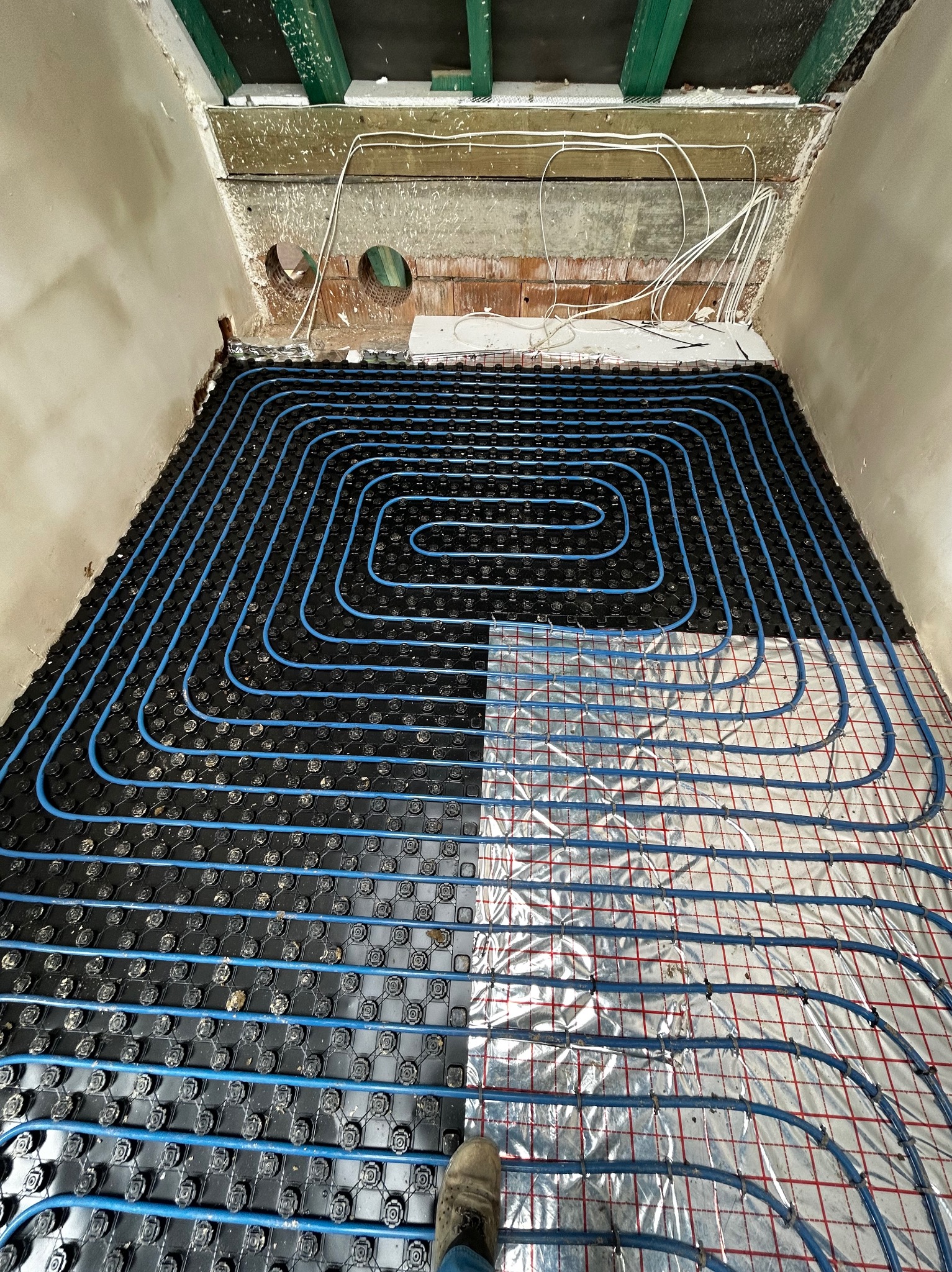 Niedokończona instalacja ogrzewania podłogowego: niebieskie rurki ułożone na czarnych mocowaniach, częściowo na izolacji z siatką, widoczne ściany w surowym stanie i drewniany strop z instalacją...