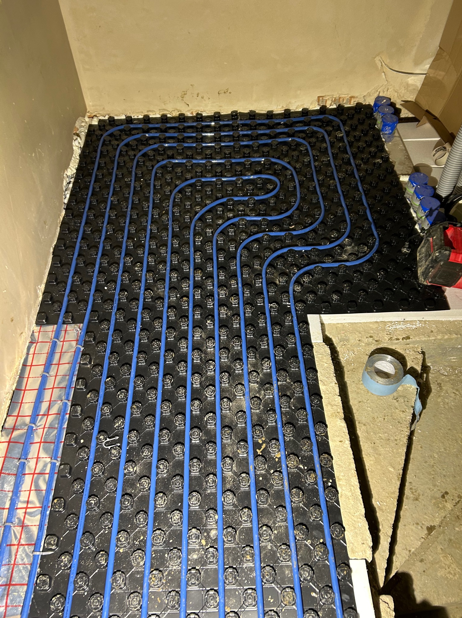 Montaż ogrzewania podłogowego w Czarnym Dunajcu: niebieskie rurki ułożone na czarnej płycie systemowej z folią aluminiową, widoczne narzędzia i materiały montażowe.