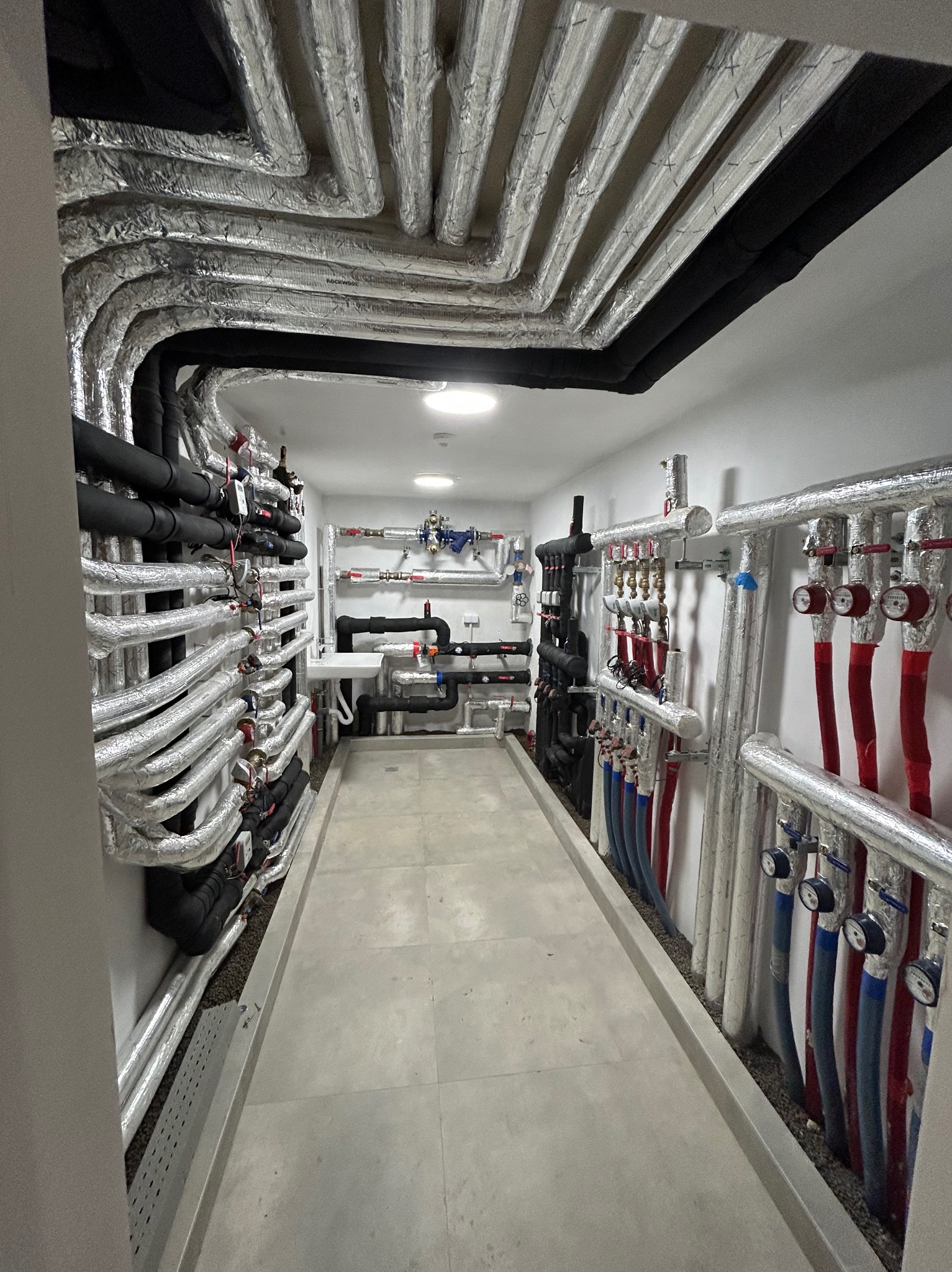 Węzeł cieplny z rzędami izolowanych rur w kolorze srebrnym i czarnym, prowadzących wzdłuż ścian i sufitu, z licznymi zaworami, manometrami i przewodami w kolorach czerwonym i niebieskim...