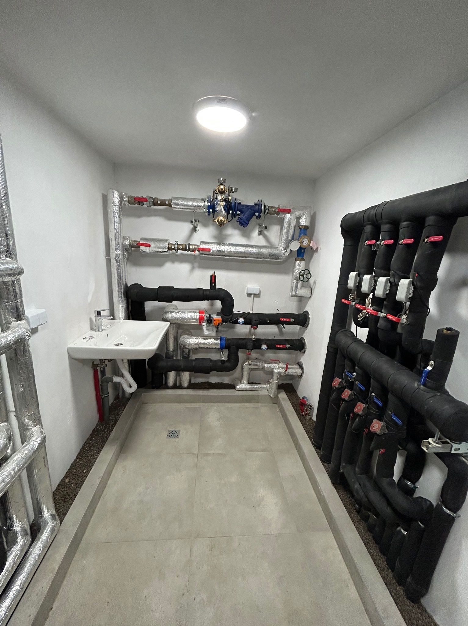Pomieszczenie techniczne z licznymi rurami instalacji grzewczej i hydraulicznej, izolowanymi i nieizolowanymi, z zaworami i armaturą, umywalką oraz betonową posadzką z odpływem.