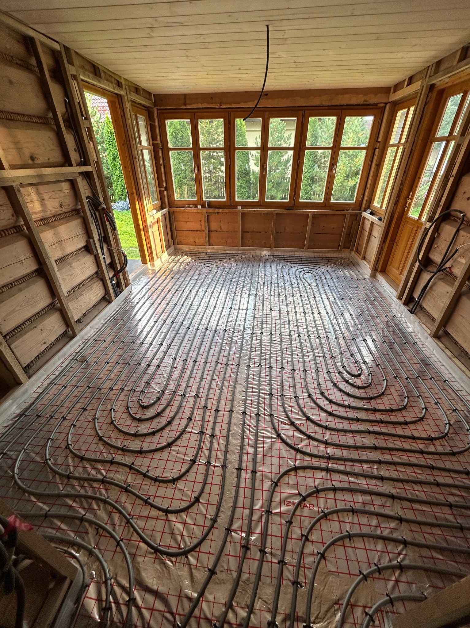 Szare rury ogrzewania podłogowego ułożone na folii z czerwoną kratką w pomieszczeniu z drewnianymi ścianami i oknami