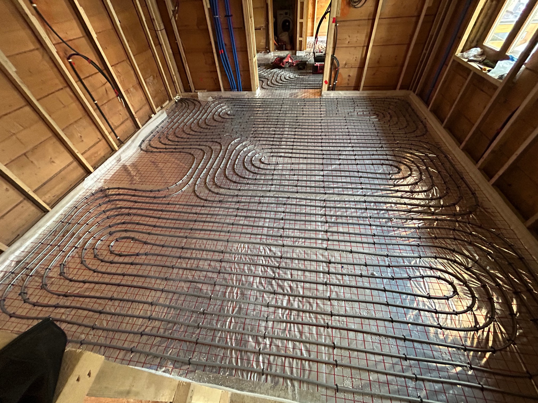 Szare rury grzewcze ułożone w pętle na metalowej siatce rozłożonej na folii izolacyjnej w trakcie montażu ogrzewania podłogowego w drewnianym budynku.