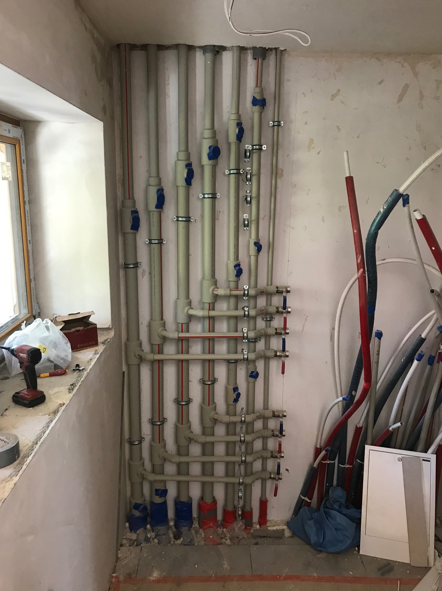 Szereg pionowych i poziomych rur instalacji wodnej z zaworami, przymocowanych do ściany w surowym stanie, widoczne narzędzia na parapecie okiennym.